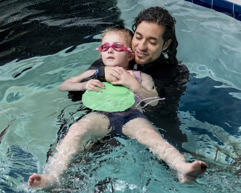 A coach holds a young swimmer steady as she practices a back float with a kickboard