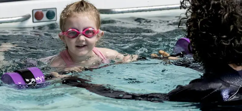 A smiling young swimmer with foam dumbbells works with her coach in the Endless Pool